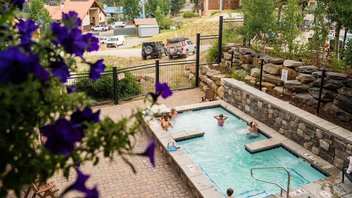 A cheerful, sunlit outdoor hot tub surrounded by lush greenery and bright flowers, with a stone wall adding rustic charm. The inviting water and relaxed atmosphere convey post-adventure leisure and social fun.