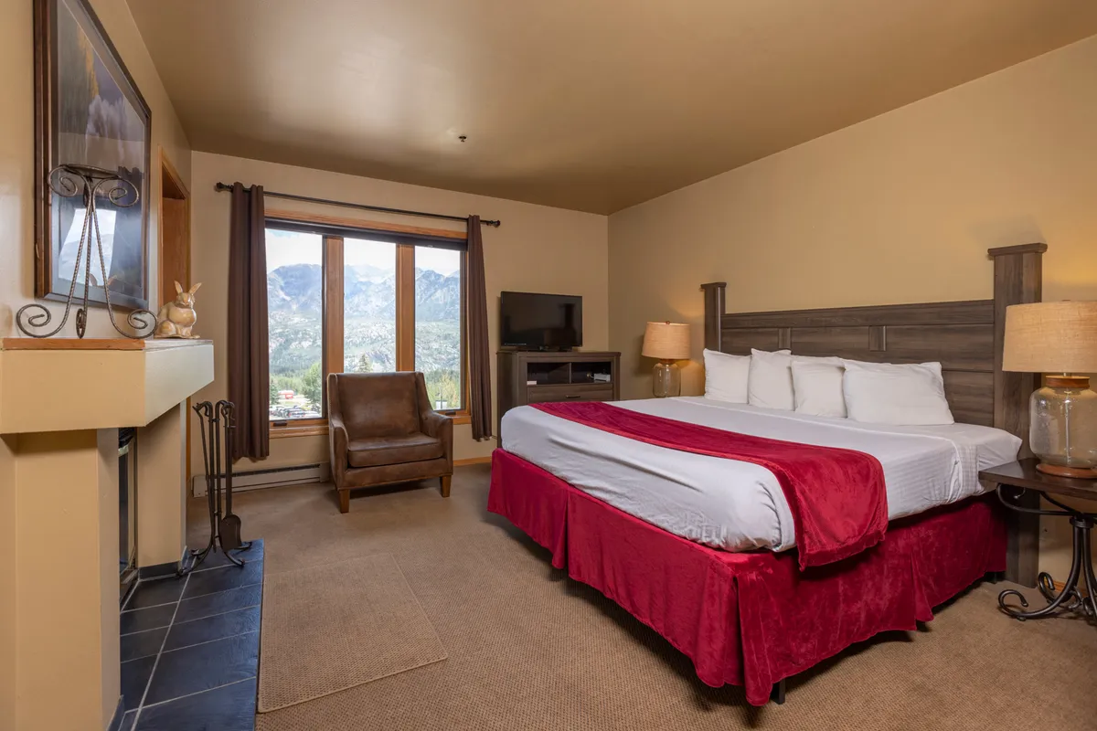 A beautifully appointed bedroom featuring a bed with a red accent, large windows offering a majestic mountain view, and a comfortable chair positioned by the fireplace. The cozy ambiance is enhanced by natural light and inviting textiles, making the room an ideal sanctuary for rest.