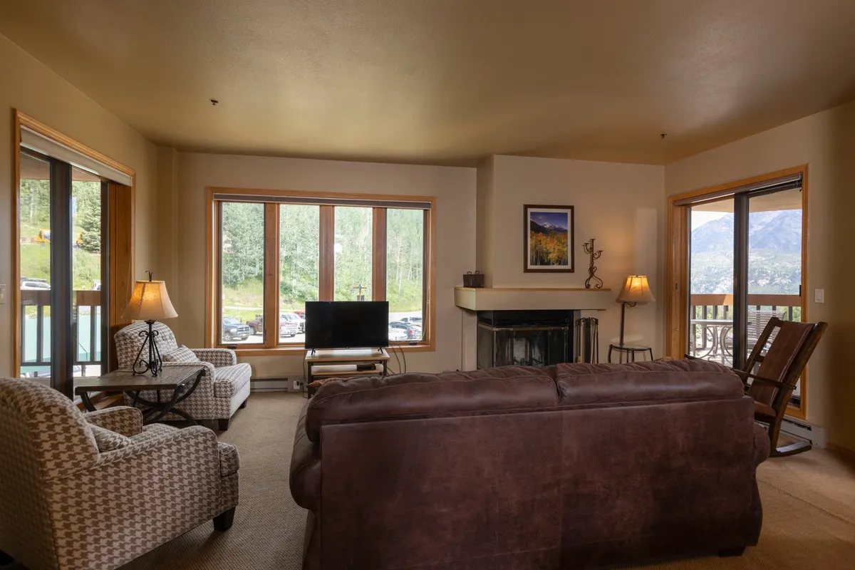A welcoming living room featuring a cozy sofa set arranged around a stone fireplace, with a television above the mantel. Expansive windows offer panoramic mountain views, and a soft rug and plush seating create an inviting atmosphere for gathering and relaxing.