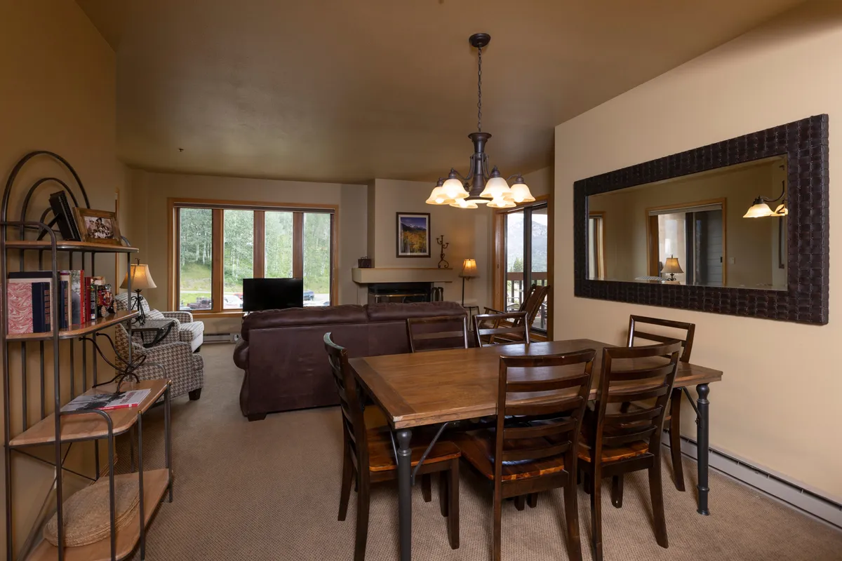 An open-plan living and dining area with a long dining table, comfortable sofa, and a fireplace. Large windows provide abundant natural light and showcase the surrounding mountain scenery. The arrangement invites group activities, conversation, and relaxation.
