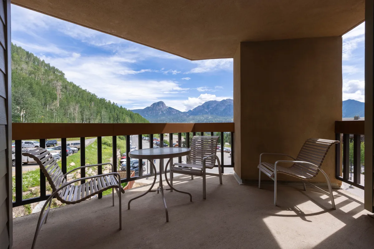 This exterior image features a balcony with outdoor seating and a table, overlooking sweeping mountain vistas. The tranquil scene invites relaxing moments, whether savoring a morning coffee, watching the sunset, or stargazing at night. The surrounding landscape is lush, and the seating area is positioned to maximize enjoyment of the view and connection to nature.