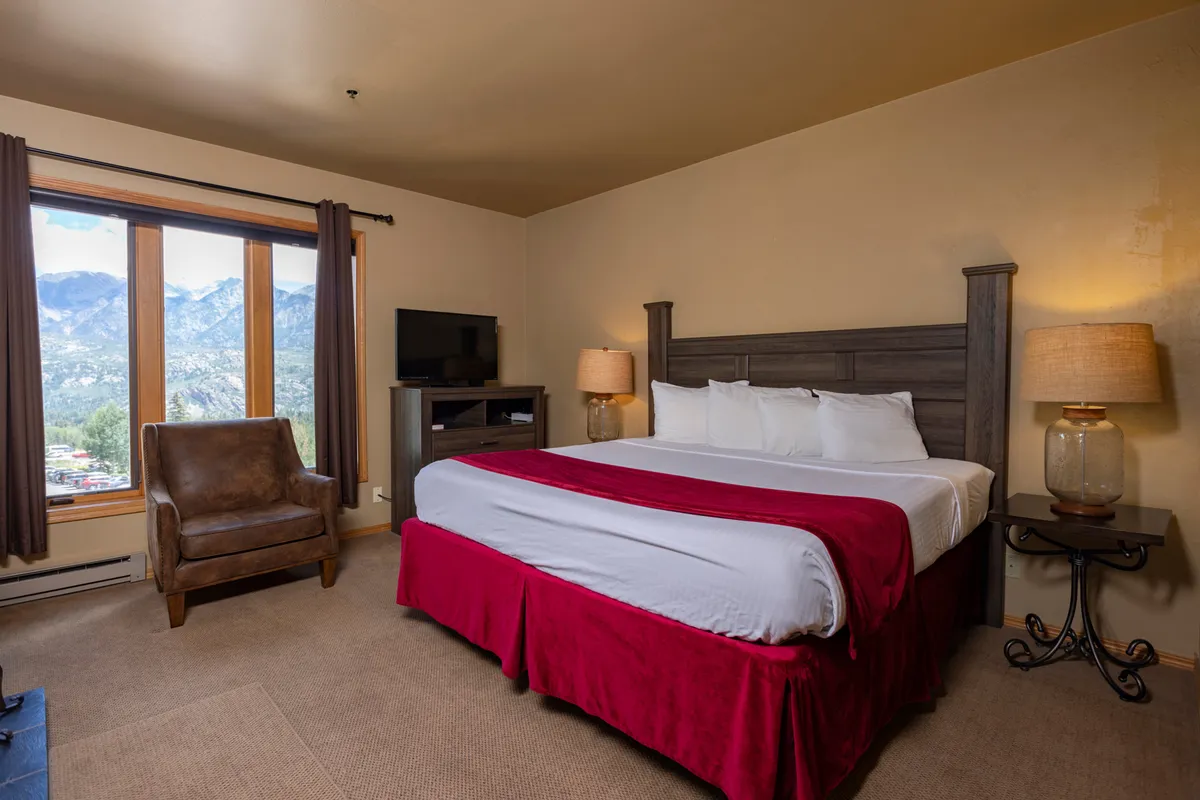 A spacious, warmly decorated bedroom with a red bed skirt, large windows framing beautiful mountain scenery, a plush armchair for reading, and a TV unit for entertainment. The space radiates comfort and peace, ideal for unwinding after a day of adventure.