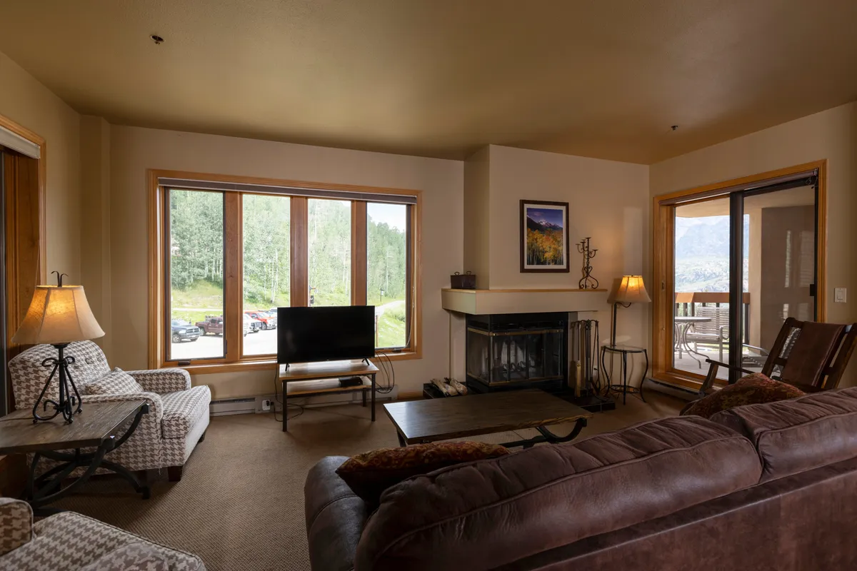 The hero image showcases a cozy living area inside a Durango condo, featuring comfortable seating arranged around a glowing wood-burning fireplace. Expansive windows bring in natural light and offer views of the changing seasons outside, inviting guests to relax and connect with the mountain setting.