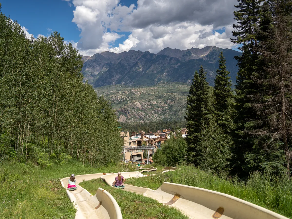In the foreground, two people enjoy sliding down a green hill surrounded by lush grass, with mountains rising dramatically in the background. The playful scene exudes a sense of summertime exploration and community enjoyment.