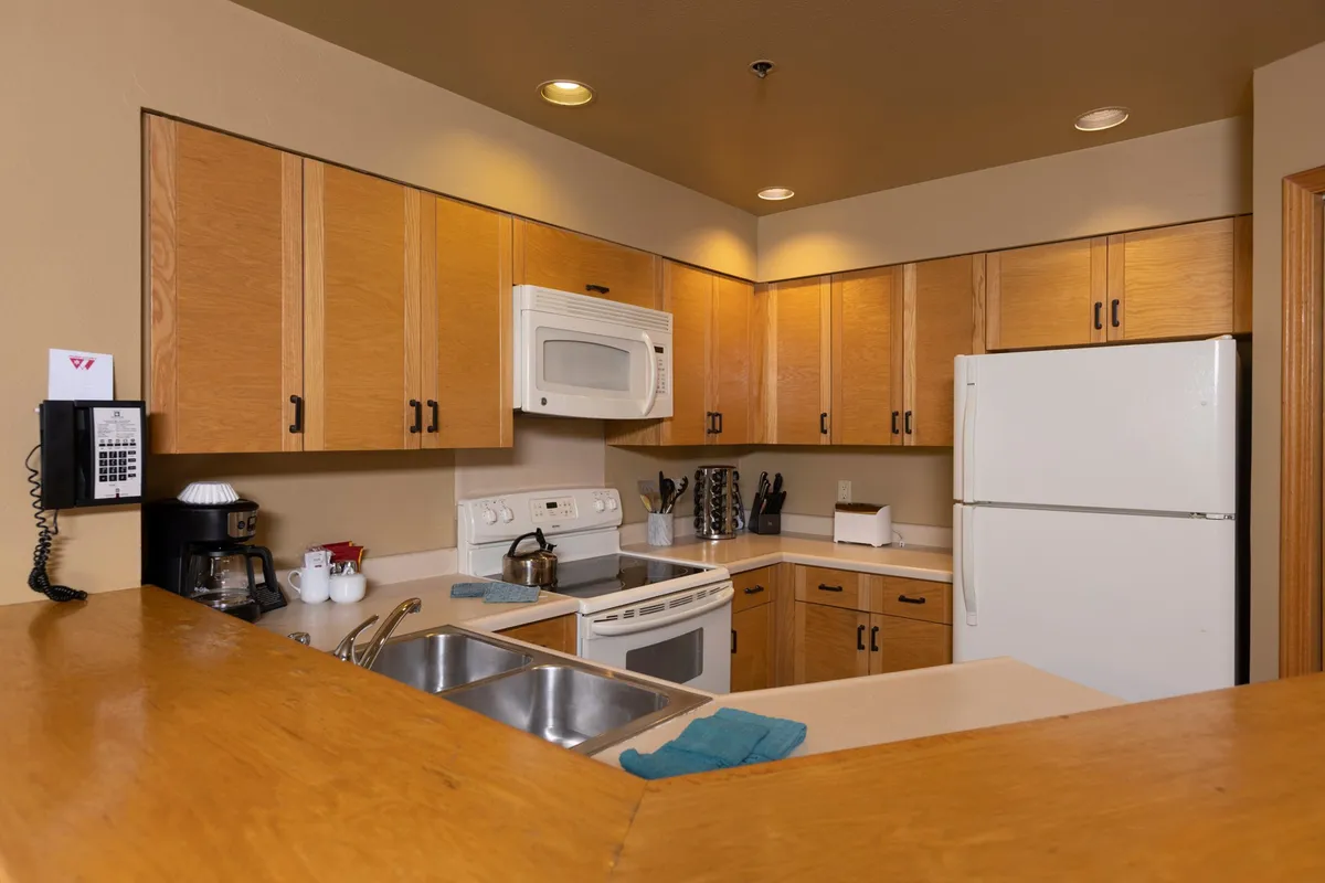 A comfortable kitchen scene featuring wooden cabinets, a wide counter for meal prep, white appliances, and a deep sink—presented in a warm, homey style. The setup feels practical and inviting, ideal for both everyday cooking and entertaining.