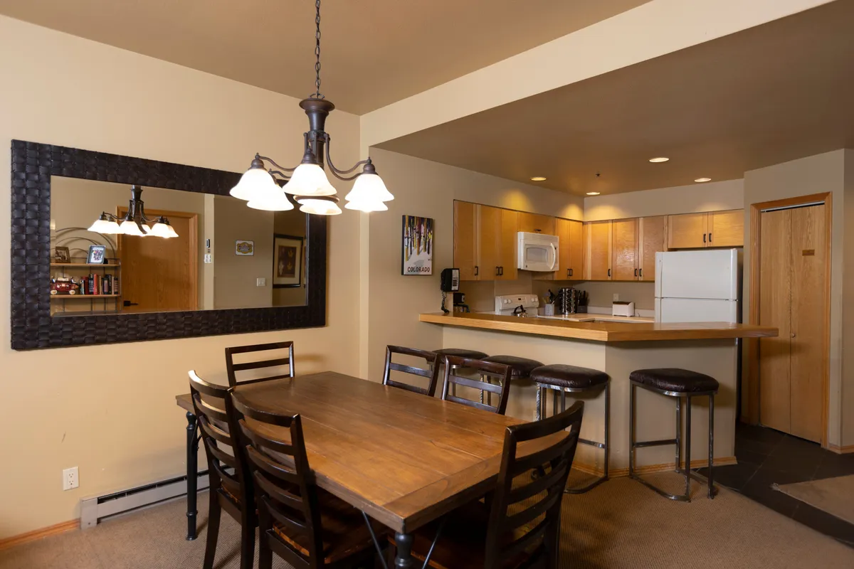 An inviting dining area centered around a large communal table, enhanced by an elegant chandelier above and a view into the adjacent kitchen bar. The setup radiates warmth and is ideal for group meals, celebrations, or intimate dinners.