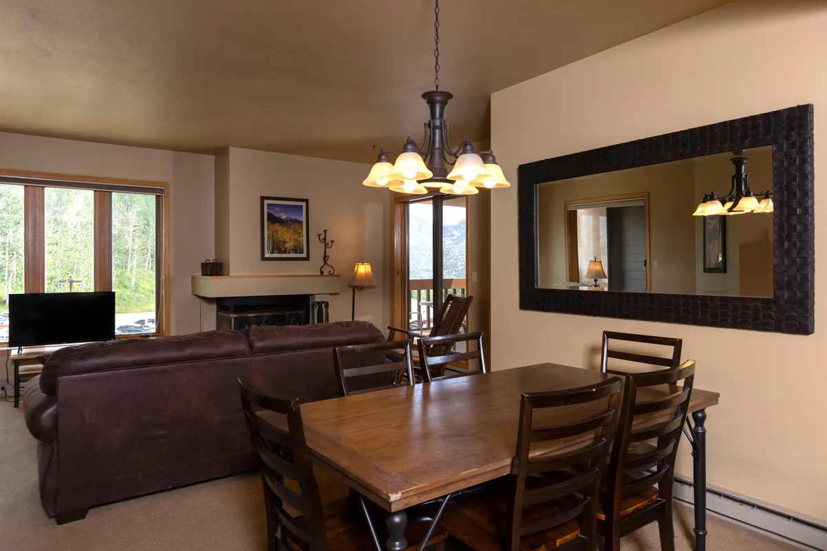 A bright, cozy space with a dining table, plush sofa, stone fireplace, and large windows. The chandelier and mirror add touches of warmth and character. The arrangement encourages storytelling and sharing after a day outdoors.