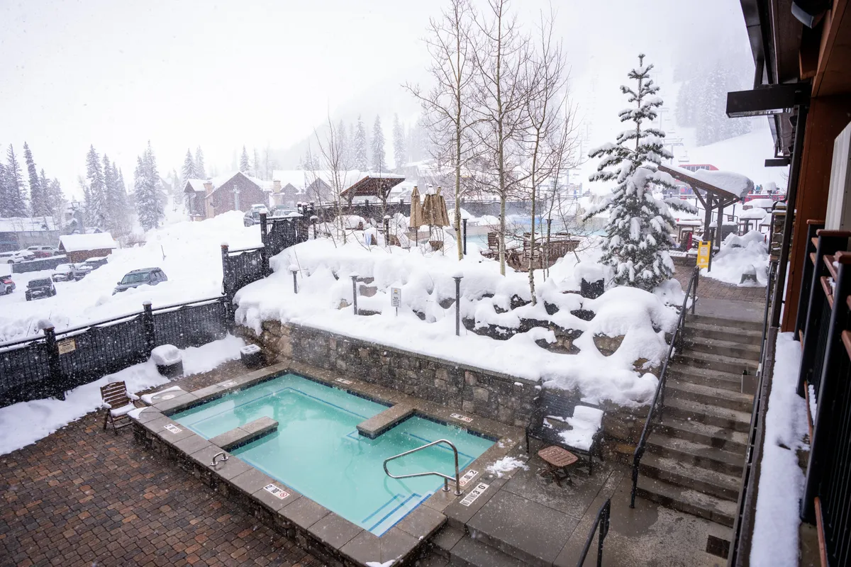 An inviting exterior hot tub sits nestled among snow-covered pine trees, with a staircase leading up to the steaming water. The landscape is blanketed in white, creating a serene winter scene perfect for relaxation and stargazing.